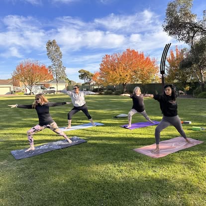 Four people practising warrior pose on the grass in a park, with autumn trees behind